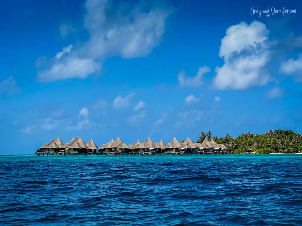 Panoramic view of a Maldivian resort with traditional thatched-roof overwater bungalows extending into the azure sea. The bungalows are aligned against a backdrop of lush tropical foliage on the island, under a vivid blue sky dotted with fluffy clouds. The rich blue of the ocean in the foreground enhances the serene and secluded setting, ideal for a peaceful and luxurious vacation destination.