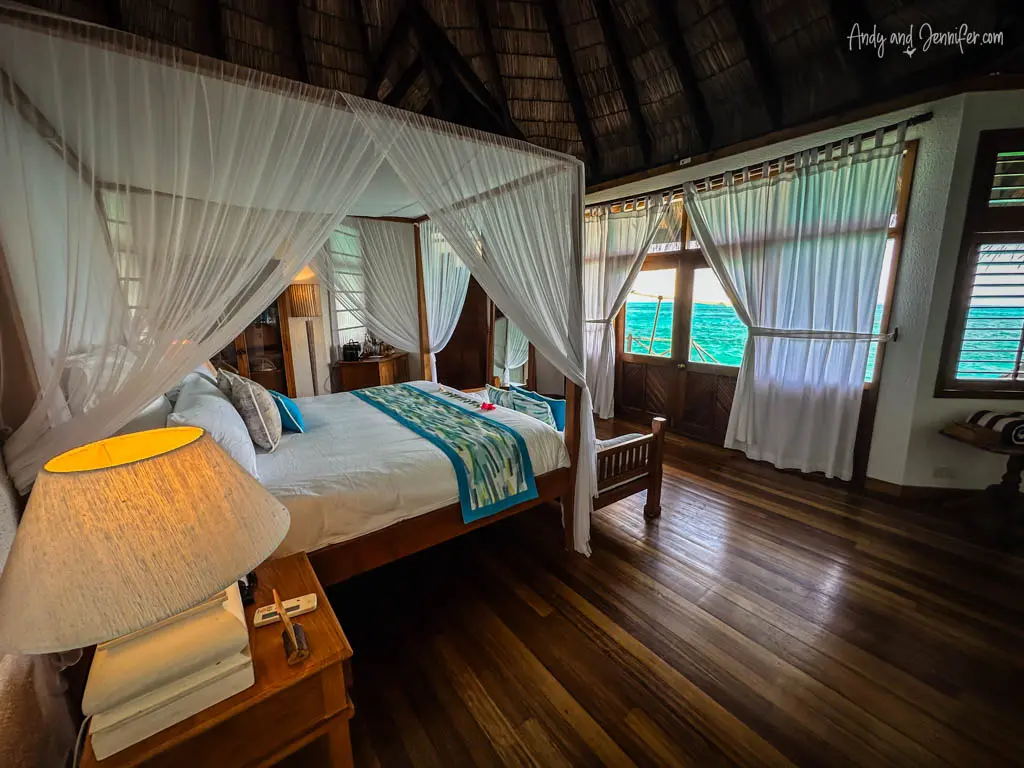 Cozy and inviting interior of an overwater bungalow in the Maldives, featuring a large bed draped with a white mosquito net. The room is constructed with rich, dark wooden floors and high ceilings, enhancing its rustic charm. Natural light floods in through windows covered with sheer curtains, offering views of the turquoise sea outside. A bedside lamp casts a warm glow, creating a peaceful, romantic atmosphere ideal for relaxation by the ocean.