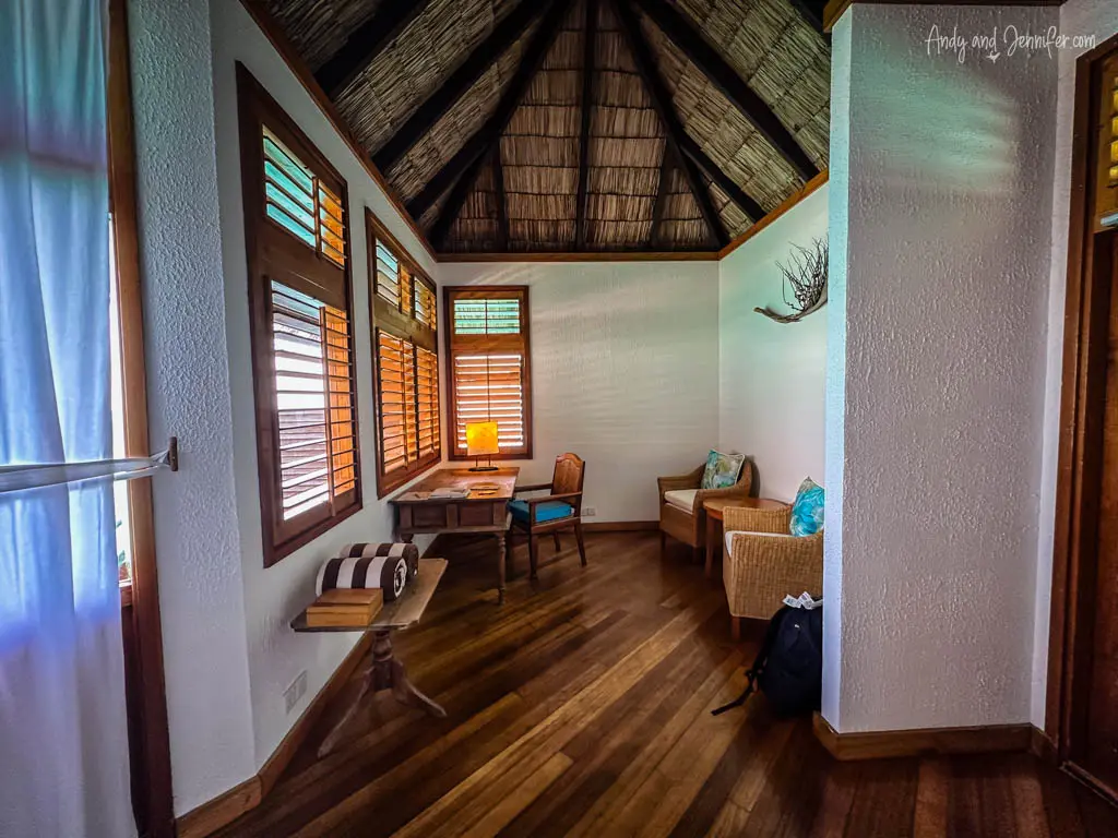 Cozy corner in a Maldivian overwater bungalow, featuring a rustic writing desk against wooden louvered windows that provide warm, ambient light. The room's traditional thatched roof and rich wooden floors enhance its tropical charm. A small seating area with wicker furniture offers a relaxing nook for reading or enjoying ocean views, ideal for peaceful, reflective moments away from resort activities.