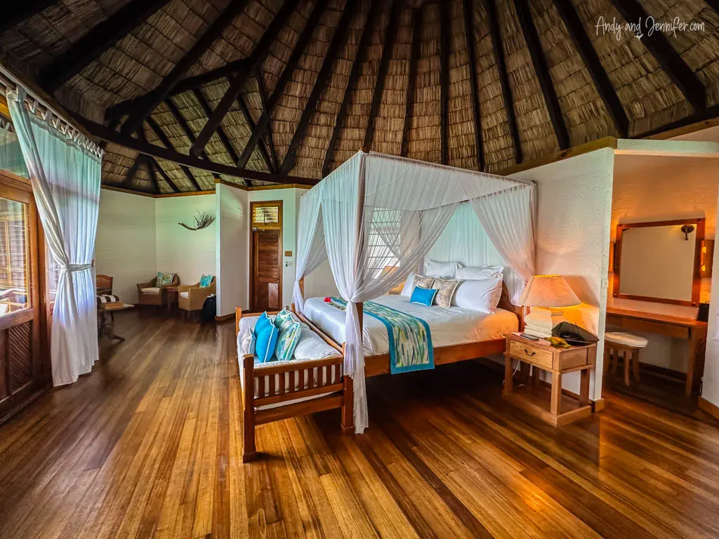 Spacious and well-appointed interior of a luxurious overwater bungalow in the Maldives, showing a large four-poster bed draped with a delicate white mosquito net. The room features high, vaulted ceilings with exposed wooden beams and rich wooden flooring, contributing to its tropical aesthetic. Ample natural light pours in through large windows dressed with sheer curtains, highlighting the comfortable seating area and elegant decor. This inviting space combines traditional Maldivian elements with modern comforts, creating an idyllic retreat overlooking the sea.