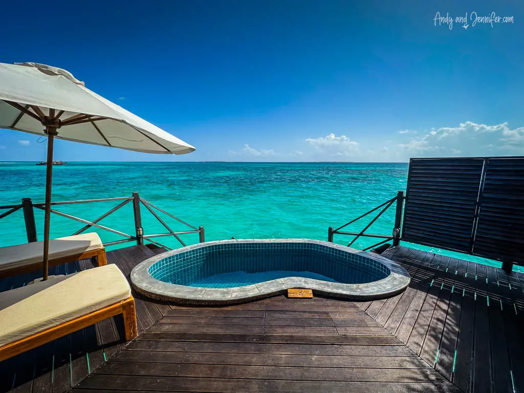Luxurious outdoor deck of an overwater bungalow in the Maldives, featuring a private jacuzzi and sun loungers under a large white umbrella. The deck is built with dark wooden planks that contrast strikingly with the vibrant turquoise waters of the ocean just beyond the safety railing. This tranquil setup offers a perfect secluded spot for relaxation and sunbathing, with panoramic sea views under the bright blue sky, ideal for a luxurious tropical getaway.