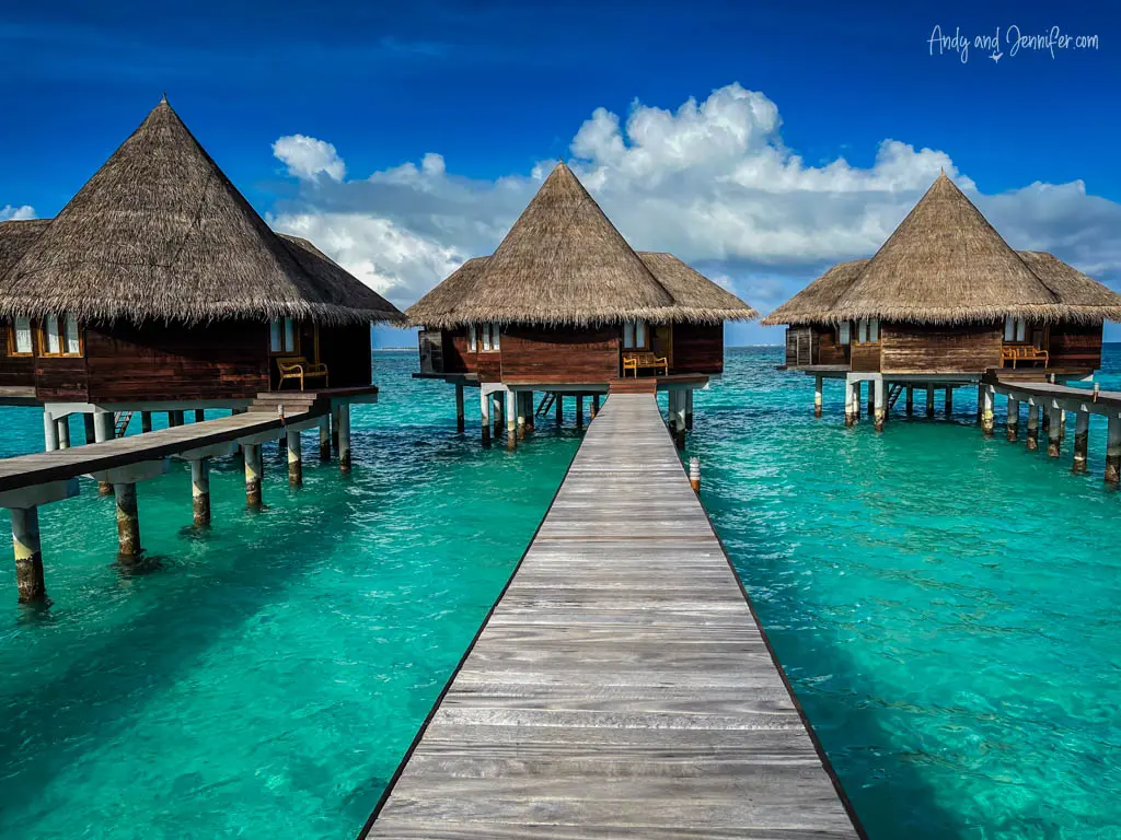 Symmetrical view down a narrow wooden jetty leading to three thatched-roof overwater bungalows in the Maldives. The turquoise sea surrounding the villas is clear, allowing visibility into the shallow water beneath. The villas are structured on stilts and feature a rustic charm with modern amenities, seen in the exterior lounge areas of each. Above, a vivid blue sky with fluffy clouds complements the tranquil and inviting oceanic landscape, perfect for a luxury vacation experience.