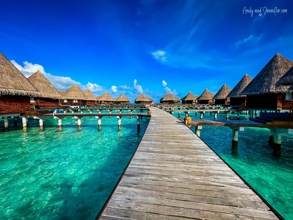 Vibrant scene of a wooden boardwalk leading to a series of luxury overwater bungalows in the Maldives. The pathway extends into the crystal-clear turquoise waters, lined with traditional thatched-roof villas on stilts. The sky is exceptionally blue with minimal clouds, enhancing the tropical and serene ambiance of this exclusive resort destination. This setting showcases the unique architecture and stunning natural beauty ideal for a luxurious seaside getaway.