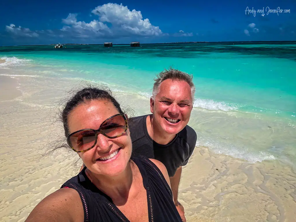 Close-up selfie of a smiling couple on a tropical beach with the stunning backdrop of the Maldives' turquoise waters and white sandy shore. The woman, wearing sunglasses, and the man, beaming with joy, capture their vacation moment under a sunny sky with fluffy clouds. In the background, the clear shallow waters blend seamlessly into the deeper blue of the ocean, where several boats are moored, enhancing the idyllic setting.