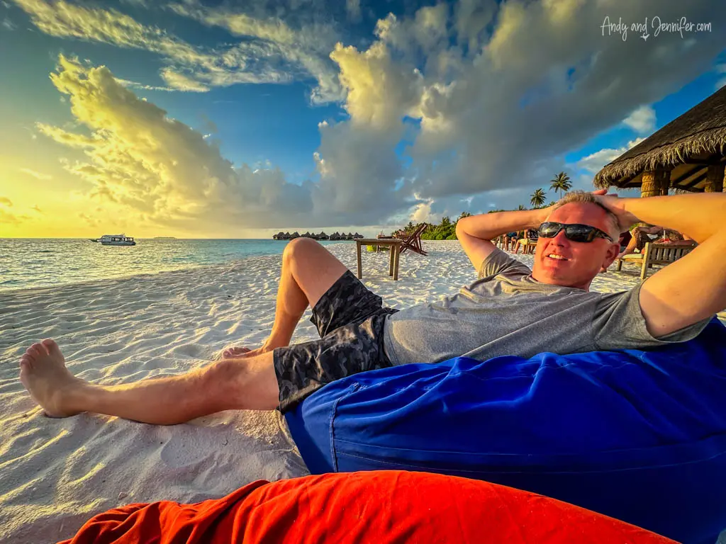Relaxed man lounging on a beach beanbag during sunset at a tropical island resort. He is wearing sunglasses and casual beach attire, with his hands behind his head, embodying a carefree vacation vibe. The background features a striking sky with dynamic clouds and a colorful sunset reflecting off the tranquil ocean, enhancing the serene and picturesque setting of this luxurious getaway.