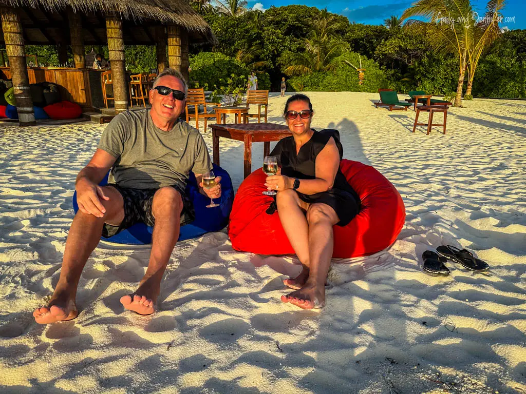 A joyful couple relaxing on vibrant red and blue beanbags on a pristine sandy beach. The man and woman are smiling, enjoying glasses of wine in a casual, serene setting, with the man wearing sunglasses and a casual gray shirt, and the woman in a black dress. Behind them, a thatched-roof beach bar and lush tropical foliage enhance the idyllic island atmosphere. This image captures a perfect vacation moment, combining relaxation with the natural beauty of a tropical beach setting.