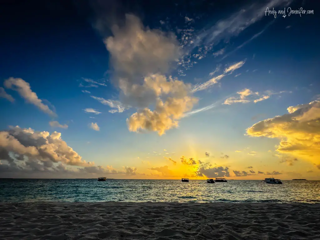 Breathtaking sunset over the ocean at a tropical beach, with the sky painted in shades of blue, orange, and yellow around scattered clouds. Sunlight pierces through the clouds, creating dramatic light rays that highlight the serene sea. A few boats are moored in the calm waters, adding to the peaceful evening seascape. The pristine sandy beach in the foreground invites viewers to experience the tranquil beauty of this idyllic setting.