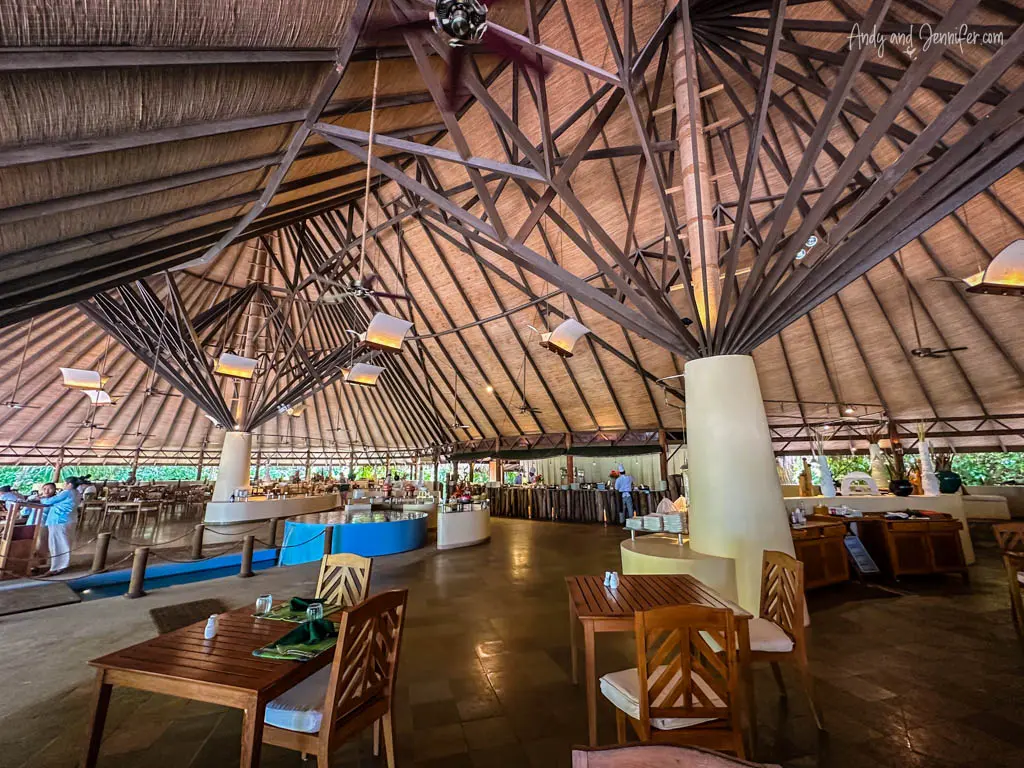 Interior of a large open-air dining hall in a Maldivian resort, featuring a high vaulted ceiling with an intricate network of wooden beams. The space is well-lit with natural lighting complemented by modern hanging lights, and it's furnished with sturdy wooden dining tables and chairs. The hall is spacious, accommodating numerous guests, with a buffet setup visible in the background, offering a variety of dishes. This inviting area blends rustic architectural elements with contemporary design, creating a welcoming atmosphere for diners.