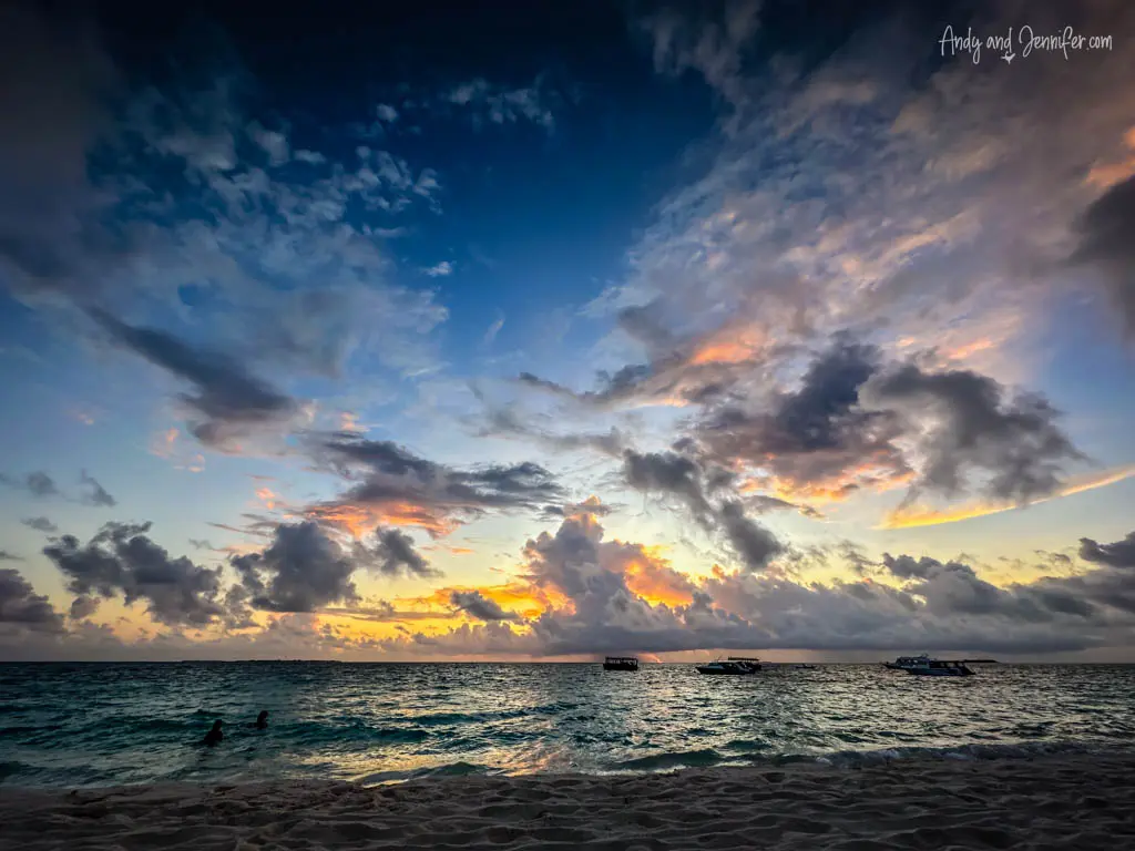 Dramatic sunset over a tranquil ocean, captured from a sandy beach. The horizon is adorned with an array of clouds, beautifully illuminated by the setting sun in hues of orange, pink, and blue. Silhouettes of small boats float peacefully in the distance, and a couple enjoys the fading light of day while swimming in the calm sea. This picturesque scene conveys a sense of peacefulness and the majestic beauty of nature during a tropical evening.