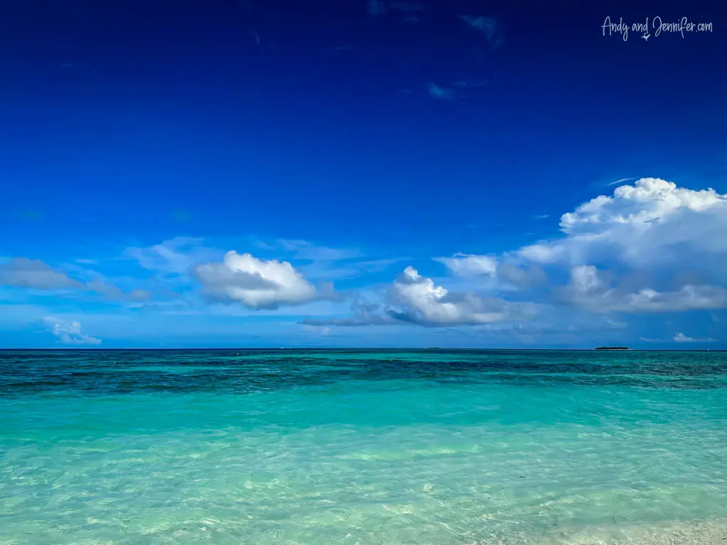 Expansive view of the Maldivian seascape with a gradient from crystal clear turquoise near the shore to deep blue in the distance. The calm sea is set against a vibrant blue sky with scattered white clouds, enhancing the serene, tropical atmosphere. This scene captures the unspoiled natural beauty and tranquility of an idyllic island paradise.