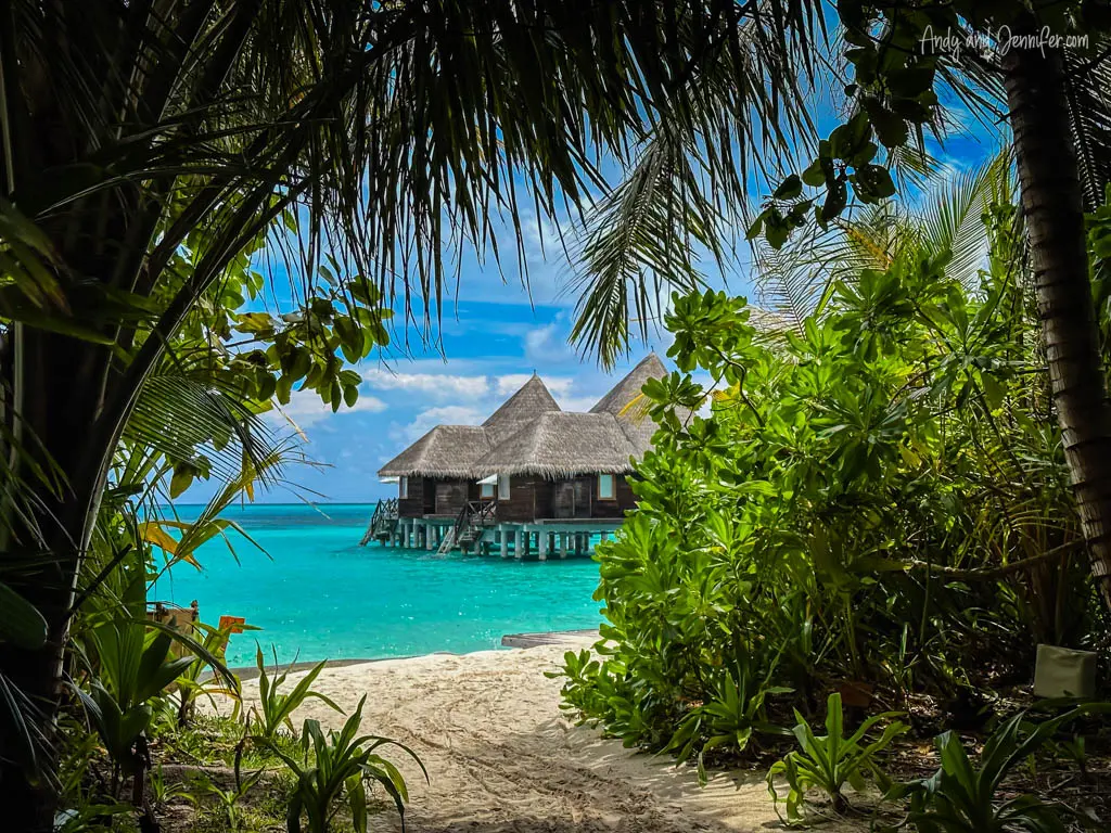 View of a secluded overwater bungalow in the Maldives, framed by lush tropical foliage and palm fronds. The pathway, covered with fine white sand, leads to the elegant wooden structure with a thatched roof, perched above the clear turquoise waters. The vibrant natural setting and the tranquility of the remote location offer a perfect escape into paradise, emphasizing privacy and the beauty of the island's coastal environment.