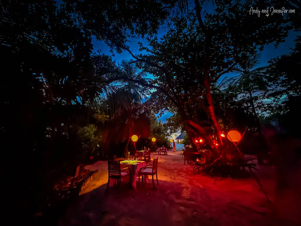 Mystical night scene at a beachside dining area in the Maldives, illuminated by soft, warm lantern light that casts a romantic glow on the surrounding sandy path and tropical foliage. A few empty tables are set up for an intimate dining experience under the stars, surrounded by the natural beauty of dense palm trees and plants, enhancing the secluded and enchanting atmosphere of this outdoor setting.