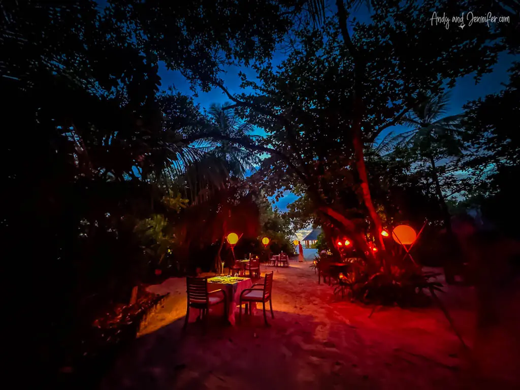 Mystical night scene at a beachside dining area in the Maldives, illuminated by soft, warm lantern light that casts a romantic glow on the surrounding sandy path and tropical foliage. 