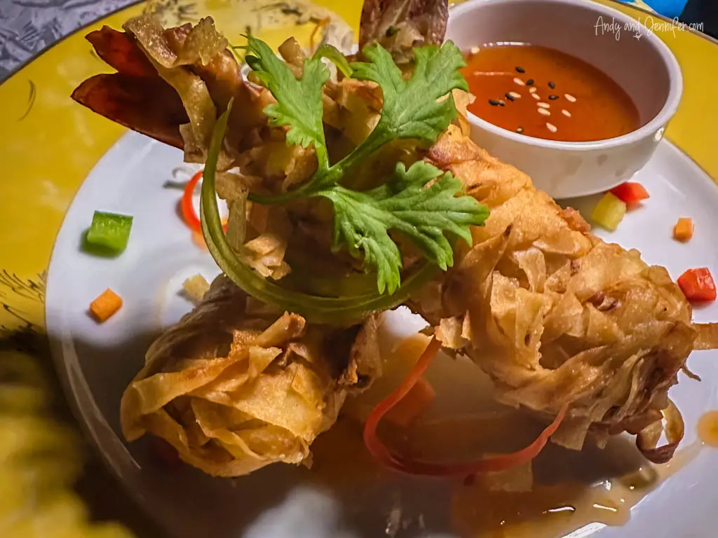 Close-up of an appetizing plate featuring crispy spring rolls artistically presented with a garnish of fresh coriander and spiral-cut vegetables. 