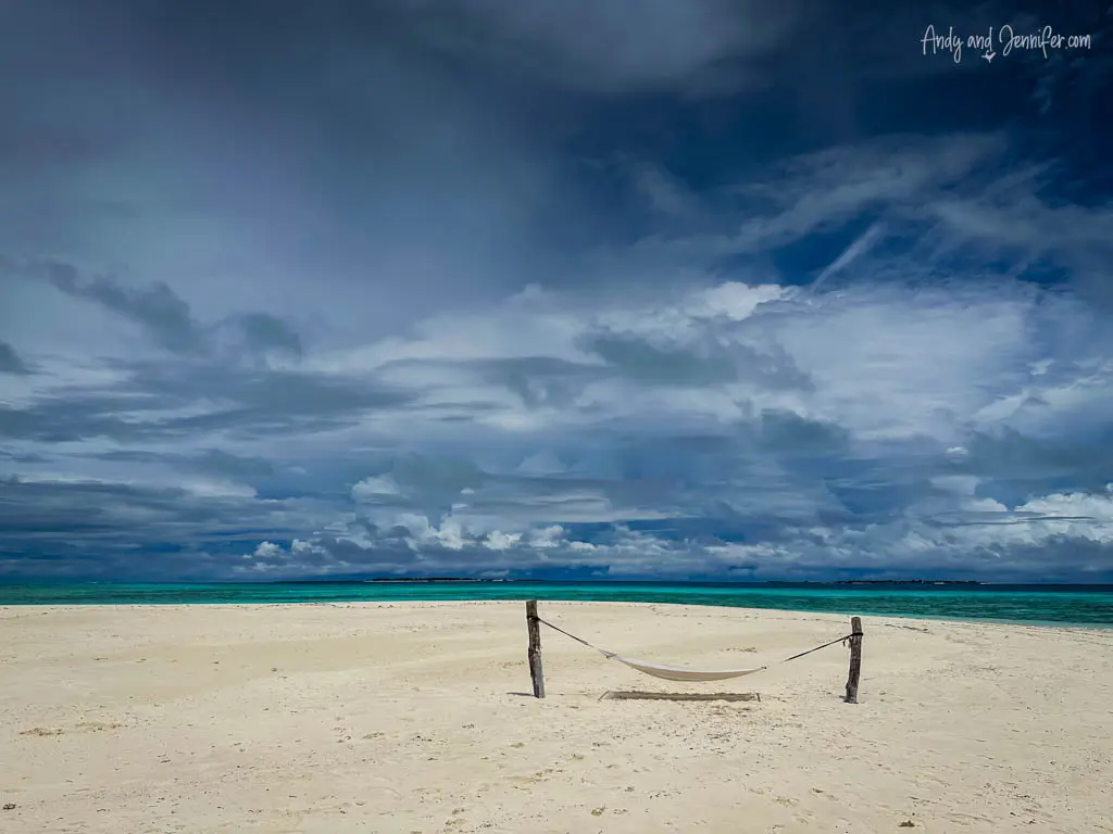 Desolate white sandy beach under a dramatic cloudy sky in the Maldives, featuring a solitary hammock tied between two wooden posts. The scene captures a stark contrast between the serene, vibrant turquoise ocean and the intense, moody blue sky, filled with dynamic cloud formations. This tranquil yet powerful natural setting evokes a sense of solitude and peace, ideal for reflection and relaxation in a remote tropical paradise.
