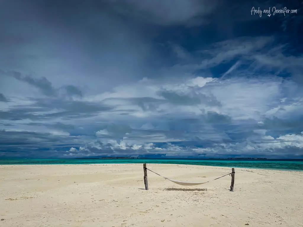 Desolate white sandy beach under a dramatic cloudy sky in the Maldives, with a solitary hammock tied between two wooden posts. The scene contrasts the serene, vibrant turquoise ocean with the intense, moody blue sky filled with dynamic clouds, evoking solitude and peace, perfect for reflection and relaxation in a remote tropical paradise.