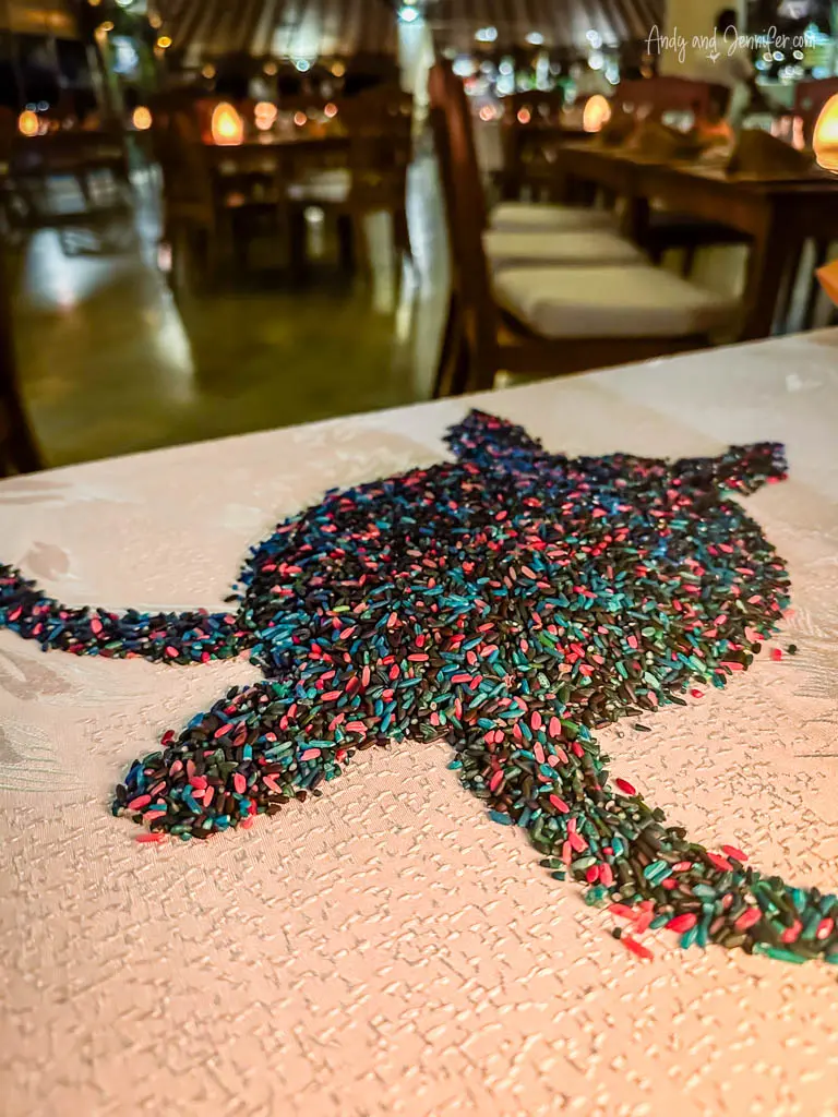 Close-up view of a creatively arranged table decoration featuring a sea turtle made from colorful rice grains, set against a textured white tablecloth. The intricate design highlights the turtle's shape and pattern, using a variety of vibrant colors to bring the artwork to life. This unique decoration is set in a dimly lit dining area, offering an atmospheric and artistic touch to the dining experience, emphasizing the resort's attention to detail and ambiance.