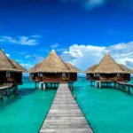 Symmetrical view of a line of luxurious overwater bungalows with thatched roofs, extending over the crystal-clear turquoise waters of the Maldives. The wooden walkway leading to each villa emphasizes the serene and pristine environment, ideal for relaxation. The vivid blue sky and fluffy white clouds above contrast with the vibrant aquatic hues, creating a postcard-perfect tropical paradise setting.