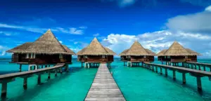Symmetrical view of a line of luxurious overwater bungalows with thatched roofs, extending over the crystal-clear turquoise waters of the Maldives. The wooden walkway leading to each villa emphasizes the serene and pristine environment, ideal for relaxation. The vivid blue sky and fluffy white clouds above contrast with the vibrant aquatic hues, creating a postcard-perfect tropical paradise setting.