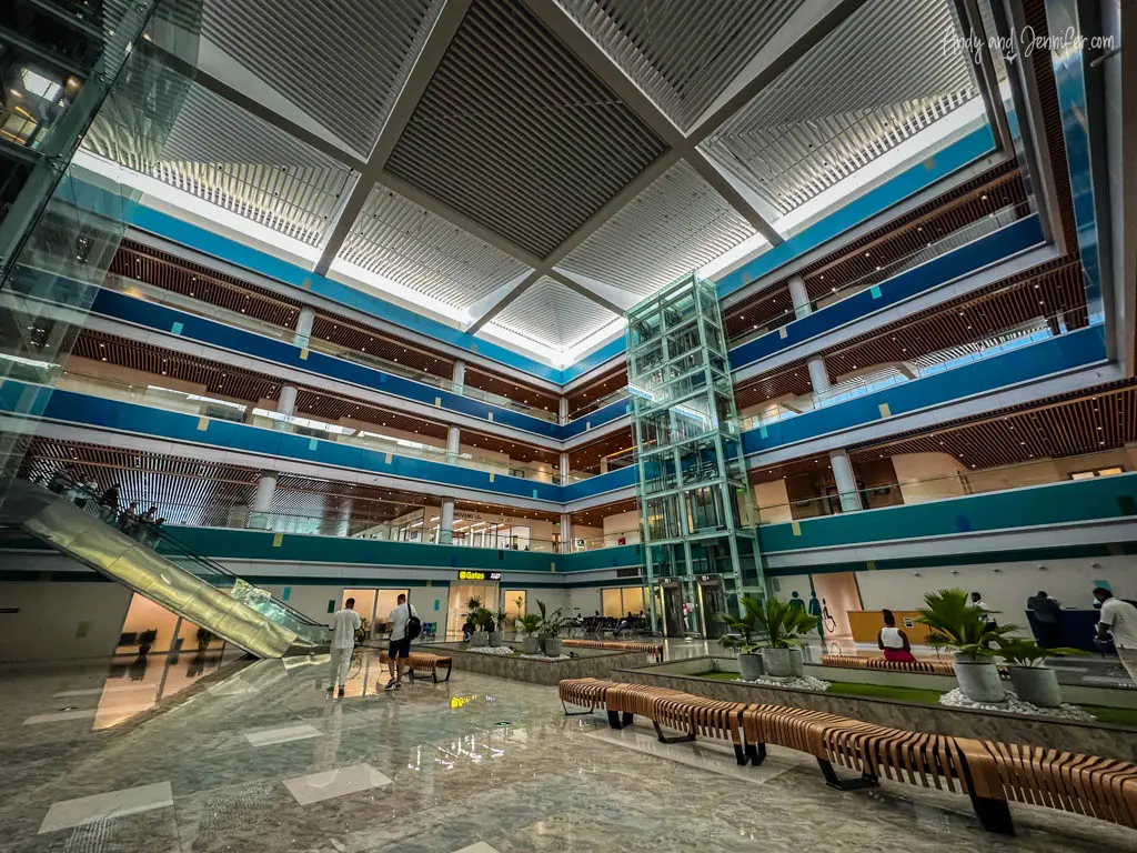 Interior of the modern seaplane terminal in Male with multiple levels visible. The design features high ceilings, extensive glass walls, and geometric patterns on the wooden balconies. The central area includes a glass elevator and a transparent escalator, enhancing the open, airy feel of the space. Seating areas with long, wooden benches and interspersed green plants create a welcoming atmosphere for travelers. The terminal is bustling with a few passengers moving about, reflecting a typical day at a busy international hub.