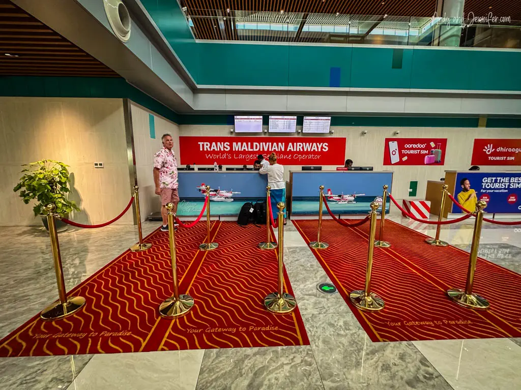 Interior of the Trans Maldivian Airways check-in area. A red carpet with wave patterns and "Your Gateway to Paradise" text welcomes travelers. Features gold stanchions, a manned check-in counter with promotional banners, a model seaplane, and Andy in a floral pink shirt, creating a lively, vacation-ready atmosphere.