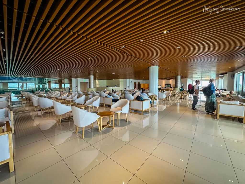 Spacious and modern Trans-Maldivian airport lounge area with contemporary design elements. The ceiling features an array of parallel wooden slats that provide a warm ambiance. The lounge is furnished with white cushioned chairs and light wood tables, arranged neatly across the glossy tiled floor, offering travelers a comfortable waiting area. Passengers are seen relaxing and some walking, contributing to the casual yet bustling atmosphere typical of an international airport lounge.
