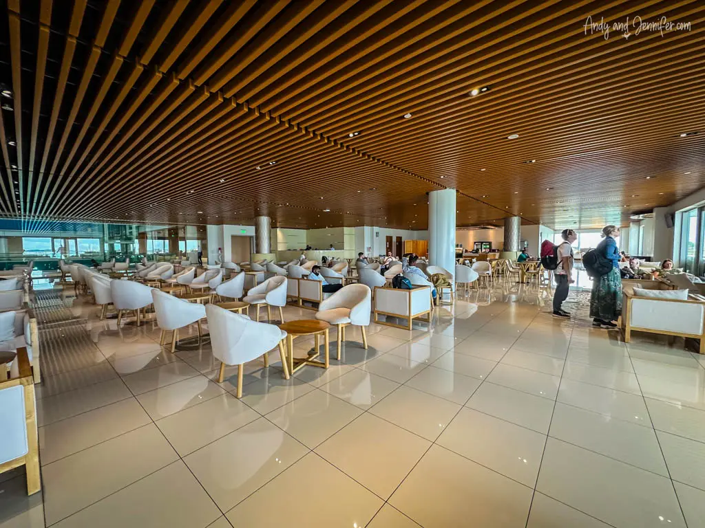 Spacious and modern Trans-Maldivian airport lounge with contemporary design elements. The ceiling features parallel wooden slats, creating a warm ambiance. The lounge has white cushioned chairs and light wood tables on a glossy tiled floor. Passengers are seen relaxing and walking, adding to the casual yet bustling atmosphere.