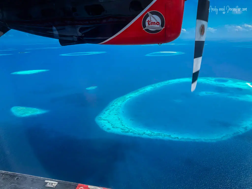 Aerial view from a seaplane over the vibrant blue waters of the Maldives showcasing several circular coral reefs forming atolls. The largest atoll is prominently visible in the center, surrounded by lighter shades of turquoise, indicating shallow waters. Part of the red and white seaplane, including the underside of the fuselage and a propeller, frames the top of the image, adding a dynamic perspective to the scenic oceanic landscape.