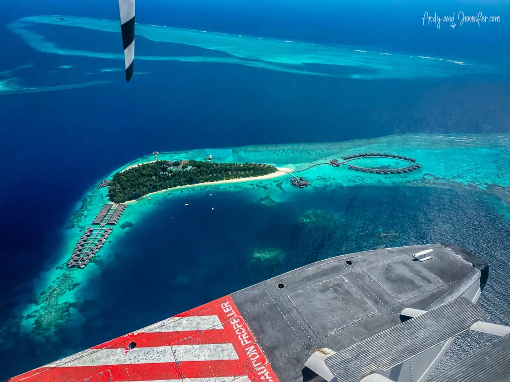Stunning aerial view from a seaplane wing over a tropical Maldivian resort island. The island is surrounded by crystal-clear turquoise waters, showcasing lush greenery, a sandy beach, and luxurious overwater bungalows arranged in a circular pattern. Part of the seaplane’s wing, painted in red and white, dominates the foreground, offering a unique perspective on the exotic destination below. The vibrant colors and scenic beauty make this image a captivating depiction of a paradise island getaway.