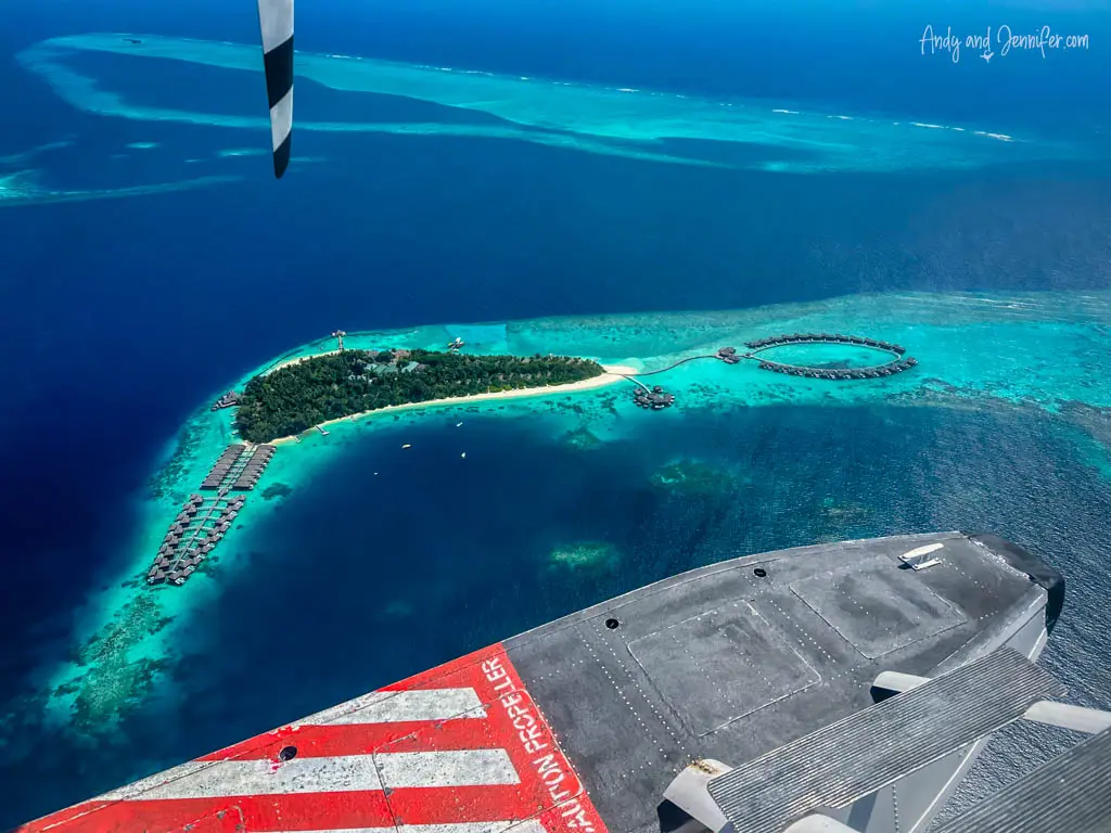 Stunning aerial view from a seaplane over a tropical Maldivian resort island. The island features lush greenery, a sandy beach, and luxurious overwater bungalows in a circular pattern, surrounded by turquoise waters. The seaplane’s red and white wing dominates the foreground, adding a unique perspective.