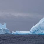 Large, towering iceberg with sharp edges floating in the ocean under a gray, overcast sky.