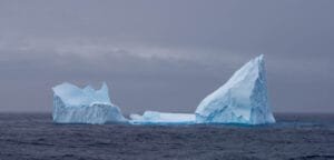 Large, towering iceberg with sharp edges floating in the ocean under a gray, overcast sky.