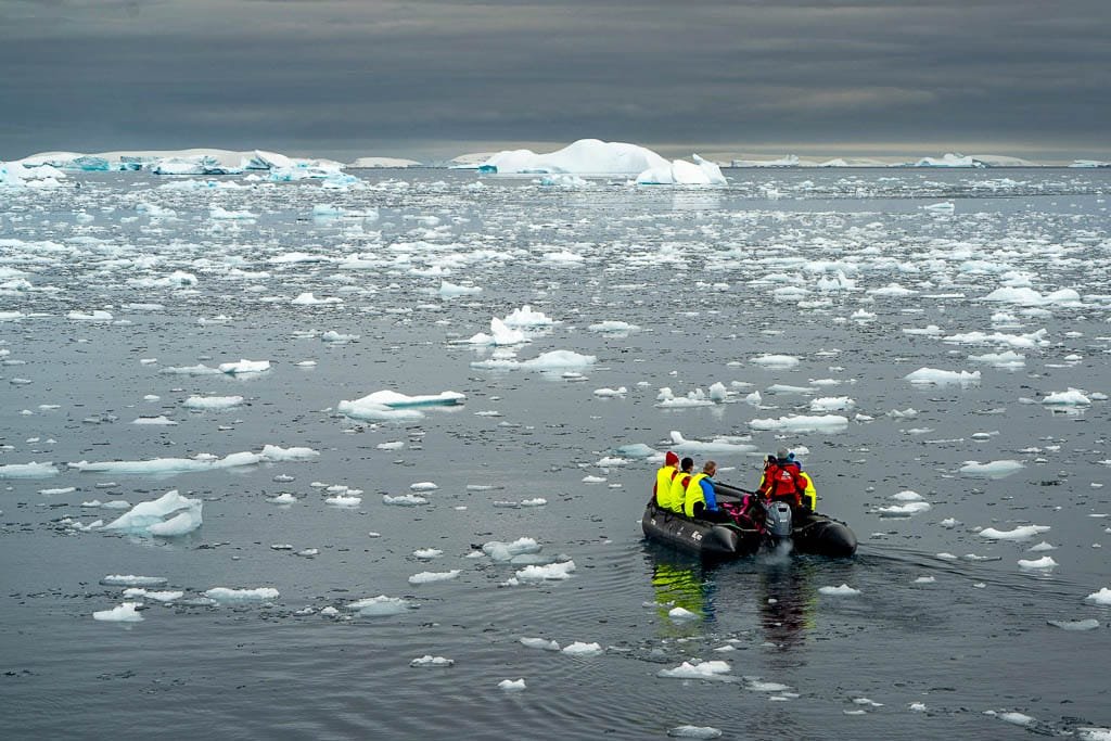  Small boat with people in yellow jackets navigating through icy waters under a cloudy sky.