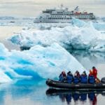 Expedition group in a small inflatable boat navigating through calm waters filled with icebergs, with a larger cruise ship in the background amidst a serene Antarctic landscape.