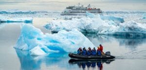 Expedition group in a small inflatable boat navigating through calm waters filled with icebergs, with a larger cruise ship in the background amidst a serene Antarctic landscape.