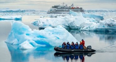 Expedition group in a small inflatable boat navigating through calm waters filled with icebergs, with a larger cruise ship in the background amidst a serene Antarctic landscape.