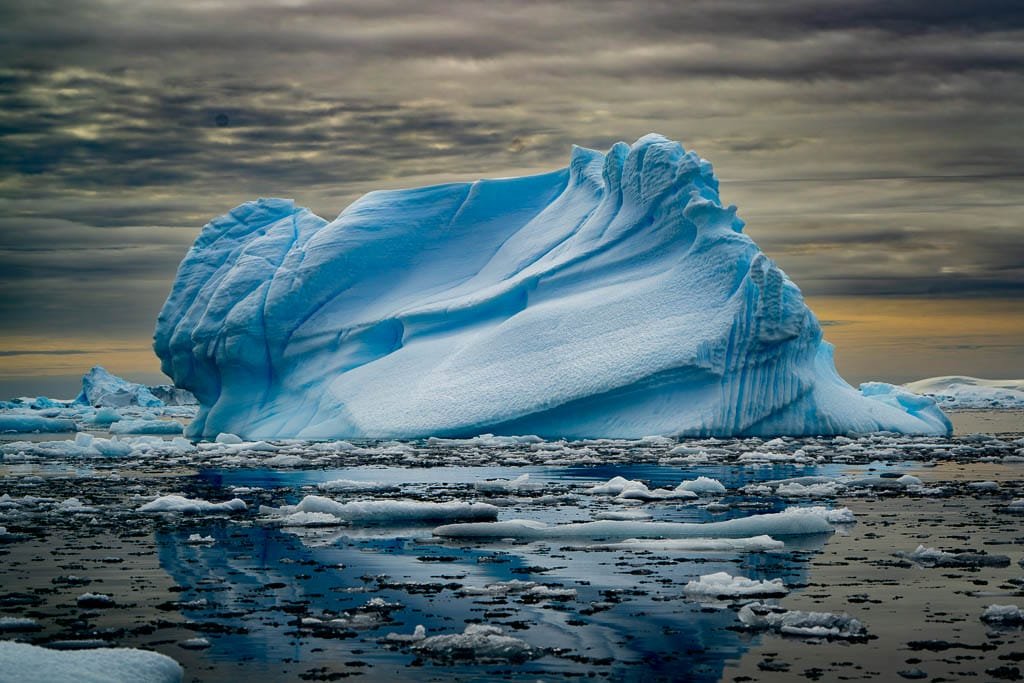 A massive iceberg with smooth, sculpted ridges floats in the dark, reflective waters of Antarctica under a dramatic, cloudy sky, tinted with hues of gray and orange from the setting sun. 