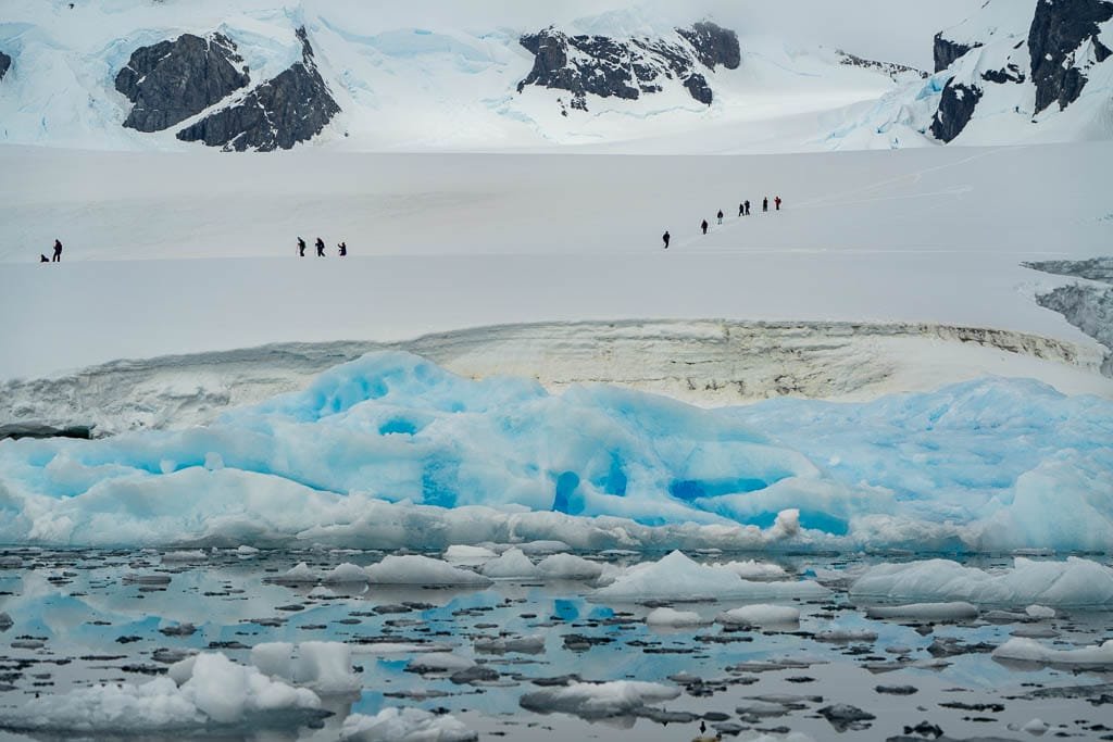  Explorers hike across a snowy Antarctic landscape with rugged mountains in the background, while blue icebergs float in the icy waters below.