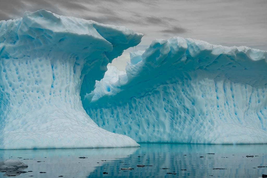 Large blue iceberg with arch formation reflecting on calm water under overcast sky.