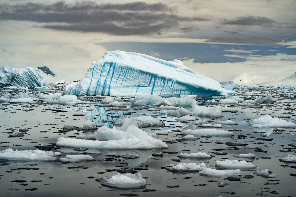 Floating blue-striped iceberg surrounded by scattered ice chunks on dark, reflective water under a cloudy sky.