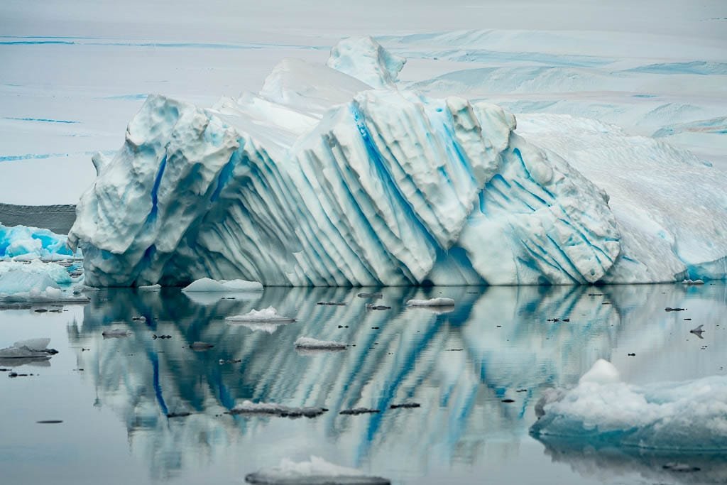 A large, uniquely shaped iceberg with blue veins and ridges reflects perfectly in the still, icy waters of the Antarctic. The iceberg's intricate structure, with sharp grooves and crevices, creates a mesmerizing pattern. Smaller floating ice pieces are scattered around, and the overall palette is dominated by icy whites and blues. 