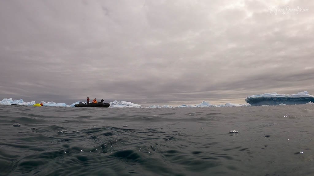 Inflatable boat floating on cold, icy water with icebergs in the distance under an overcast sky. 