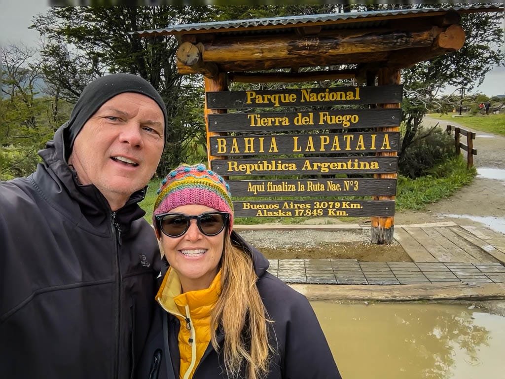Smiling couple in winter jackets posing in front of the "Bahía Lapataia" sign at Tierra del Fuego National Park, marking the end of Argentina's Ruta Nacional 3.