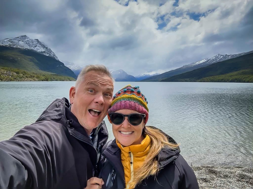 Happy couple in winter jackets posing by a serene lake with snow-capped mountains and a cloudy sky in the background. 