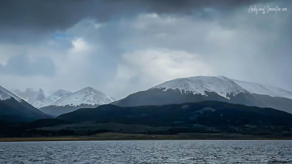 Dramatic view of snow-covered mountains under dark, cloudy skies, with a calm body of water in the foreground.