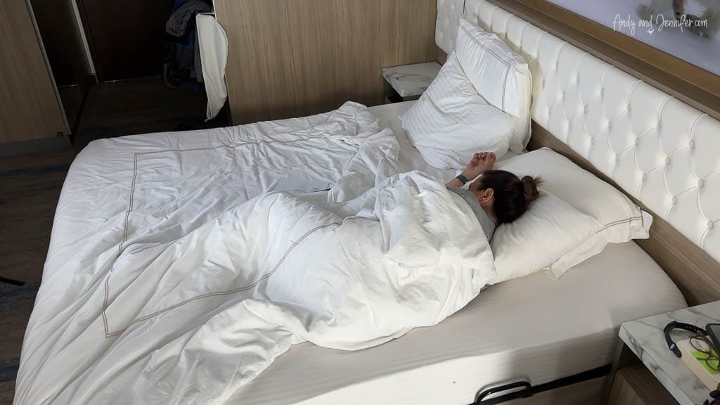 Person sleeping in a cozy bed with white sheets, wearing an eye mask in a well-lit room.