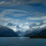 Snow-capped mountains and a glacier towering over a calm sea with a ship in the distance.