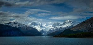 Snow-capped mountains and a glacier towering over a calm sea with a ship in the distance.