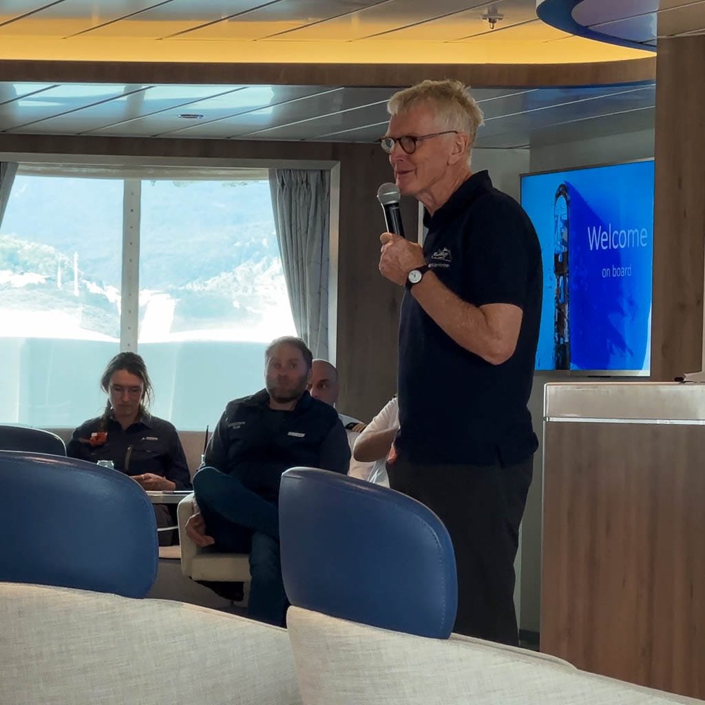 Greg Mortimer holding a microphone giving a presentation on a ship with "Welcome on board" displayed on a screen behind him. Passengers are seated and listening, with large windows showing an ocean view.