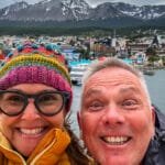 Smiling couple in winter jackets with Ushuaia's port and snow-capped mountains in the background.